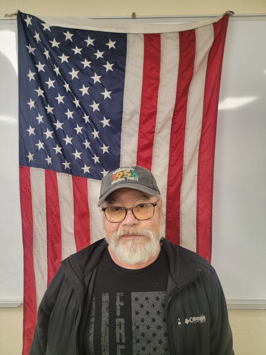 A man with glasses and a cap stands in front of an American flag.