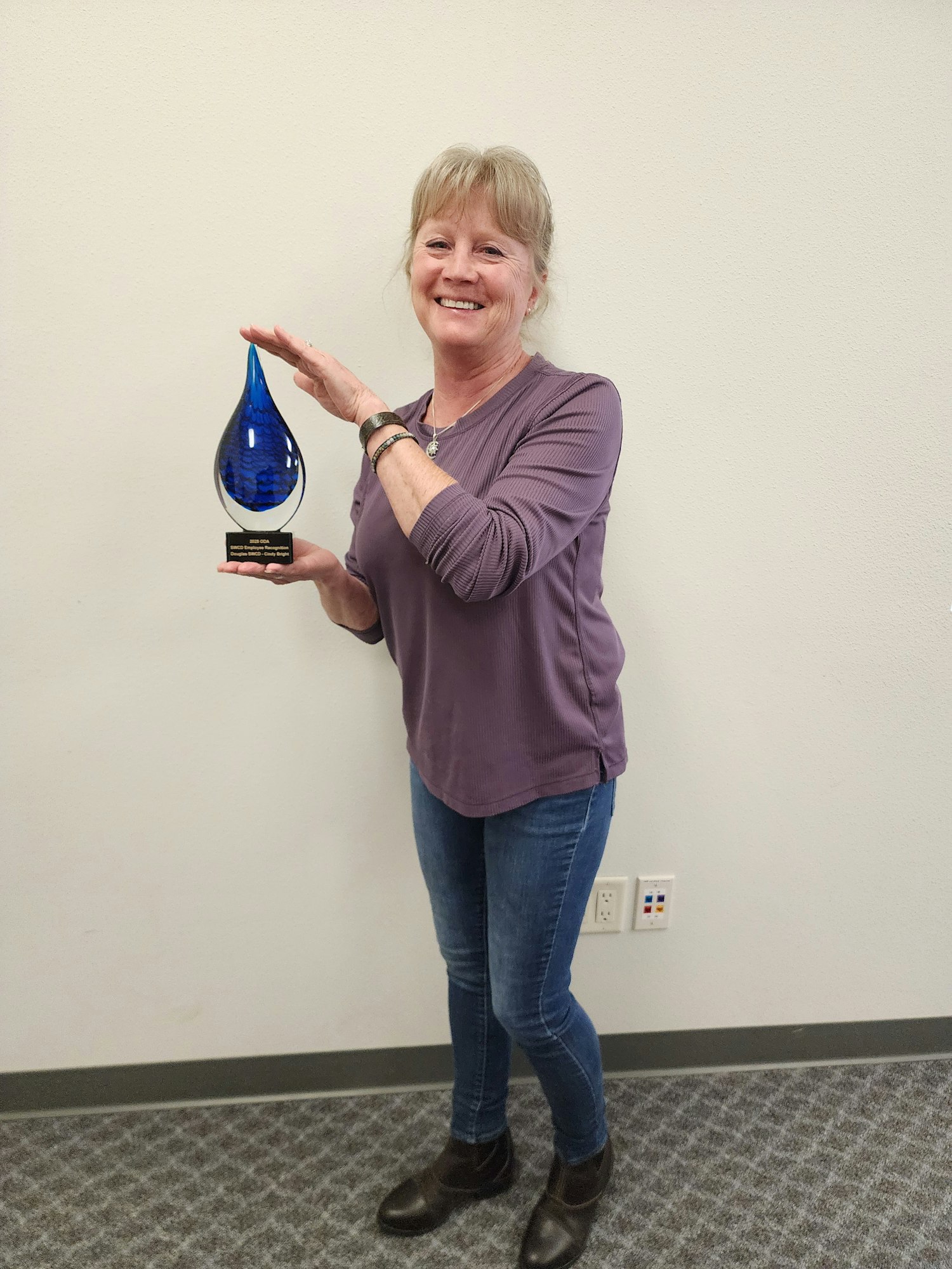 A woman holding a blue teardrop-shaped award, smiling, wearing a purple shirt and jeans.