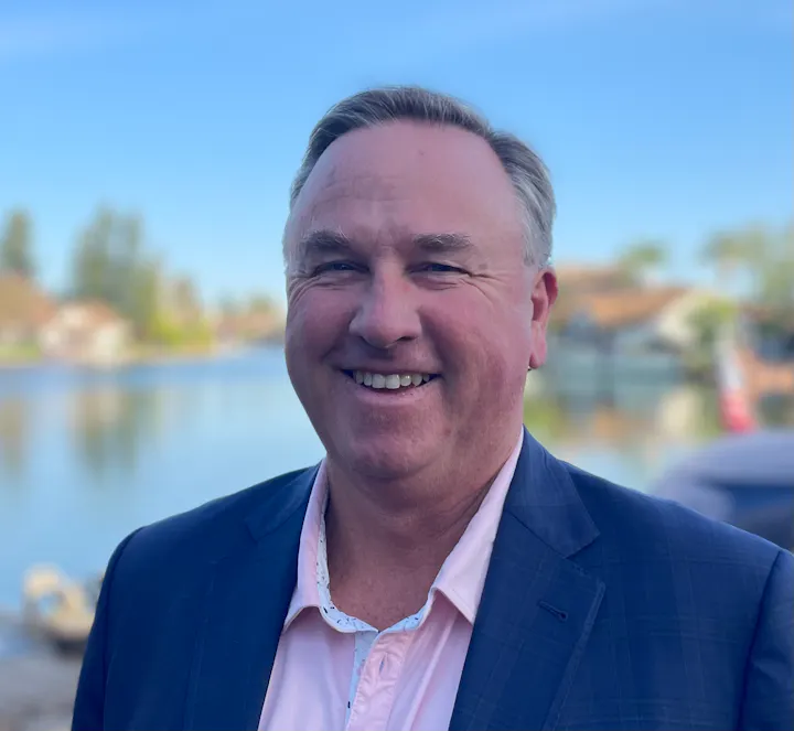 A smiling man in a suit stands by a lake on a sunny day, with houses and trees in the background.