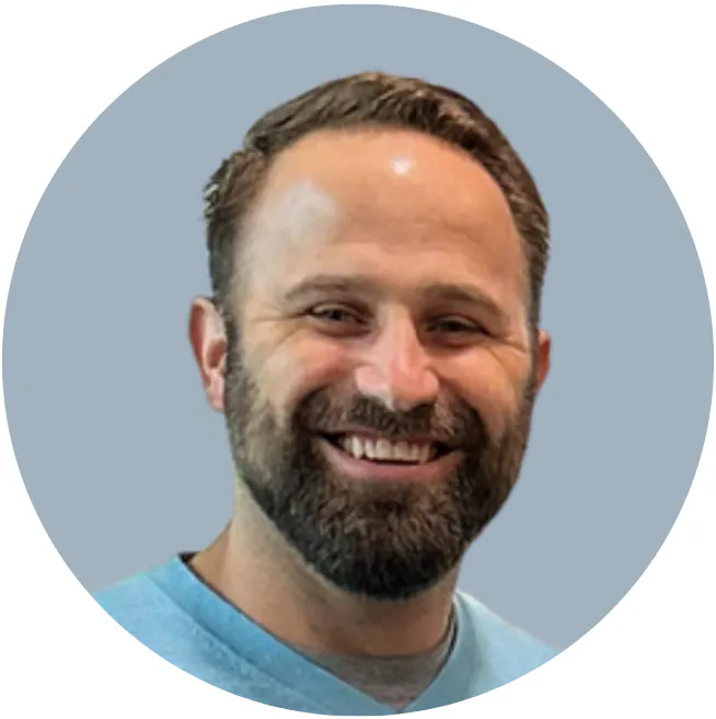 The image shows a man with a beard smiling, wearing a light blue sweater, set against a gray background.