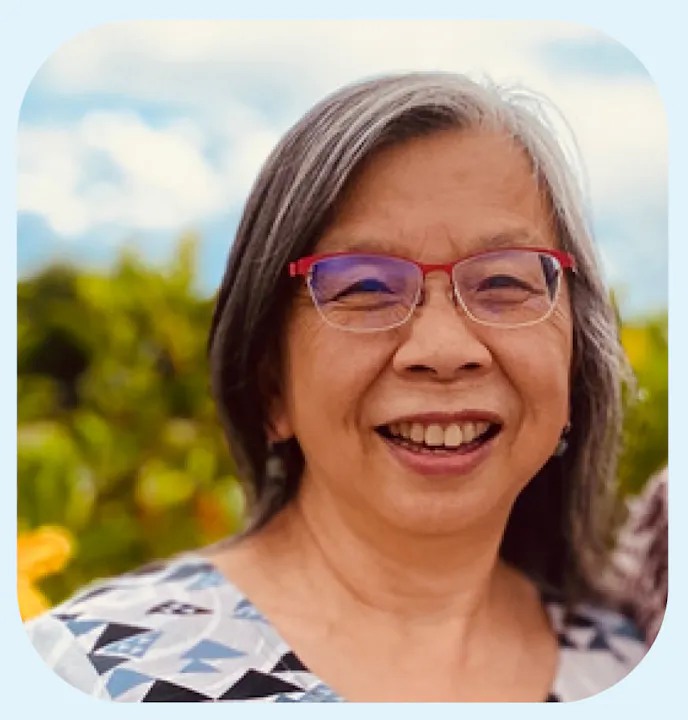 A smiling woman with glasses and gray hair, wearing a patterned top, appears happy in a natural outdoor setting.