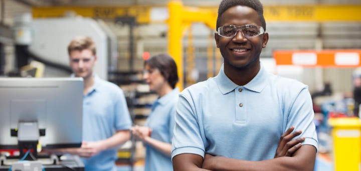 A group of people with safety glasses in an industrial setting, smiling and posing for the camera.