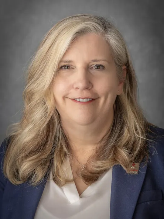 The image features a woman with long, wavy blonde hair, smiling and wearing a blazer over a white top.
