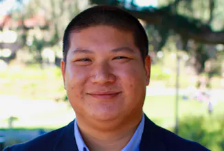 The image shows a smiling man in a suit with a blurred outdoor background.