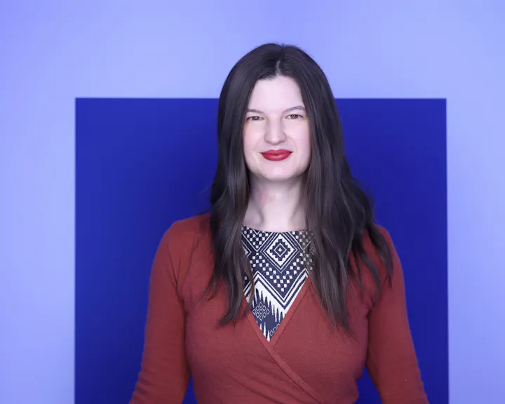 The image features a woman with long, dark hair wearing a patterned dress and a rust-colored cardigan, set against a blue backdrop.