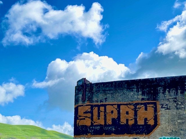 Graffiti saying "SUPAH" on a concrete wall, blue sky with clouds, green hill in the background.