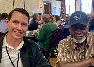 Two people seated at a table in a conference room, smiling at the camera. One is wearing a cap.