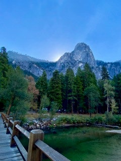A wooden bridge leads to a forest with a tall, rocky mountain in the background under a clear blue sky.