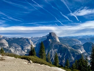 A mountain landscape with Half Dome, pine trees, and a vibrant blue sky with wispy clouds.