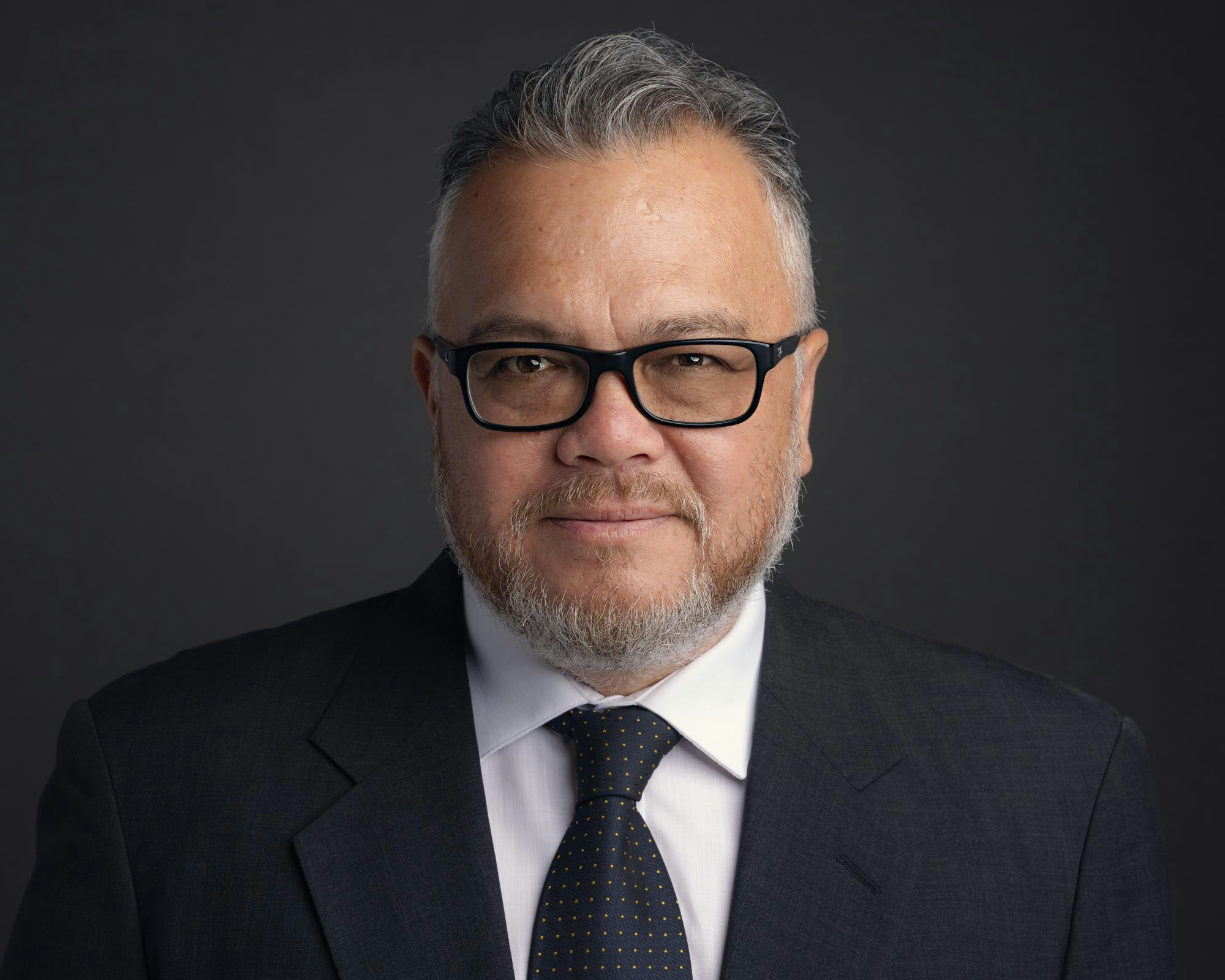 A bearded man wearing glasses and a suit with a polka dot tie against a dark background.