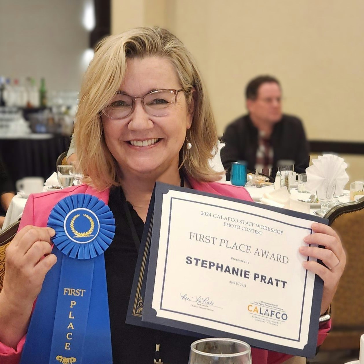 A smiling woman holding a blue ribbon and a certificate for a first-place award at a photo contest.