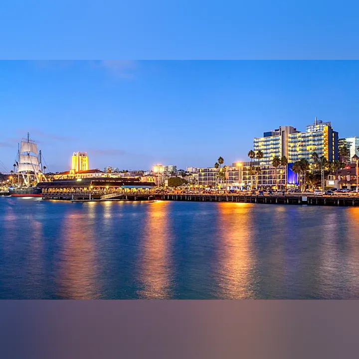 The image shows a coastal cityscape at dusk, featuring a tall ship, waterfront buildings, and a shimmering sea.