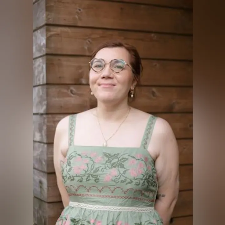 A smiling person wearing a floral dress and glasses, standing against a wooden background.
