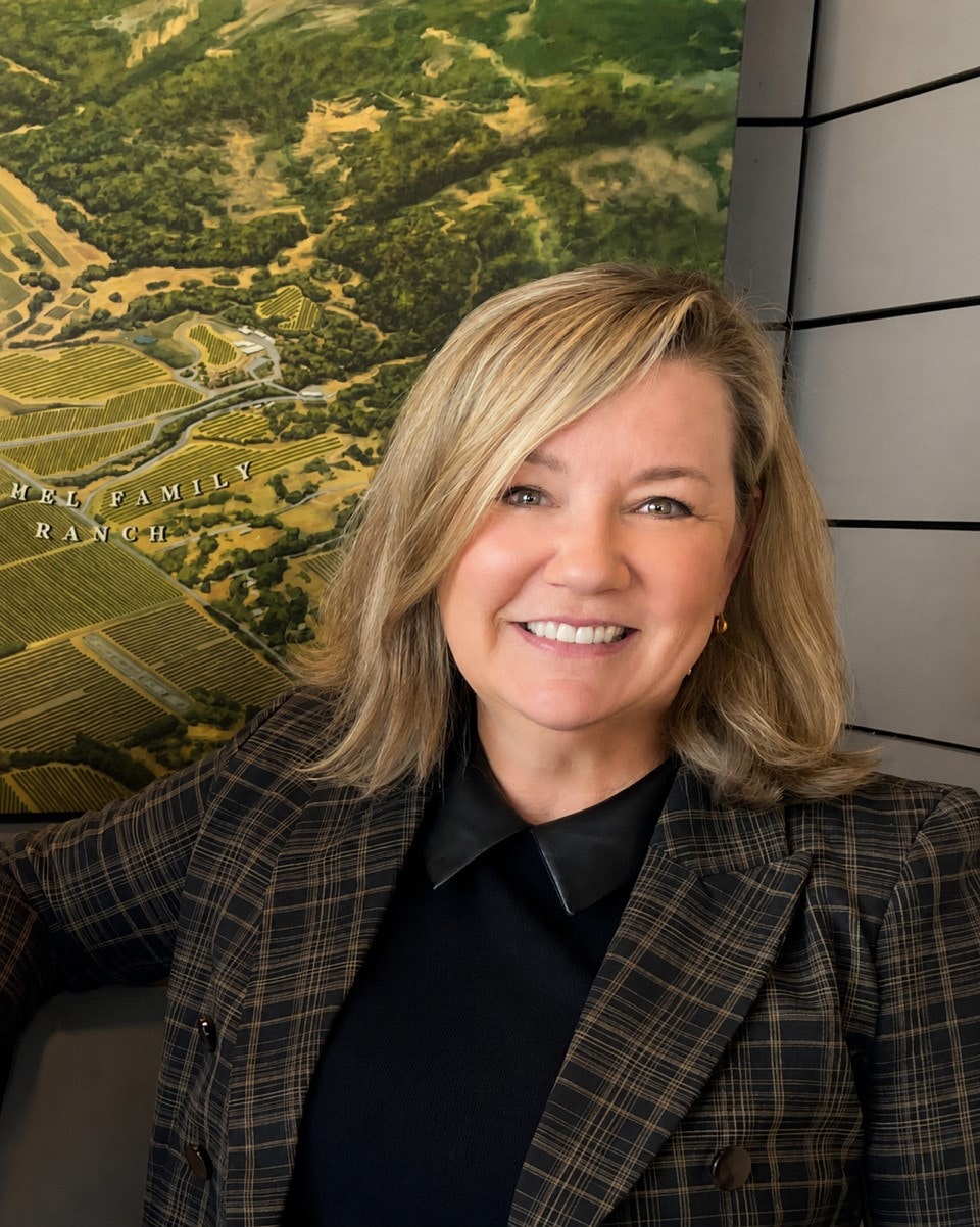 A smiling woman with blonde hair is standing in front of a large aerial image of a vineyard and ranch setting.
