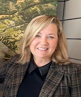 A person smiling in front of a large wall map, wearing a plaid blazer and black shirt.
