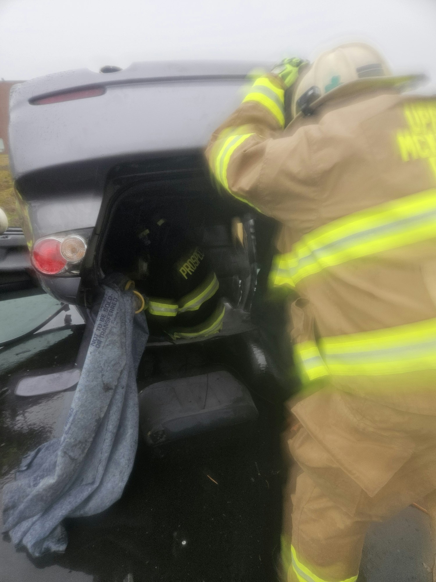 A firefighter in gear leans over a vehicle trunk, possibly during a rescue operation or inspection.
