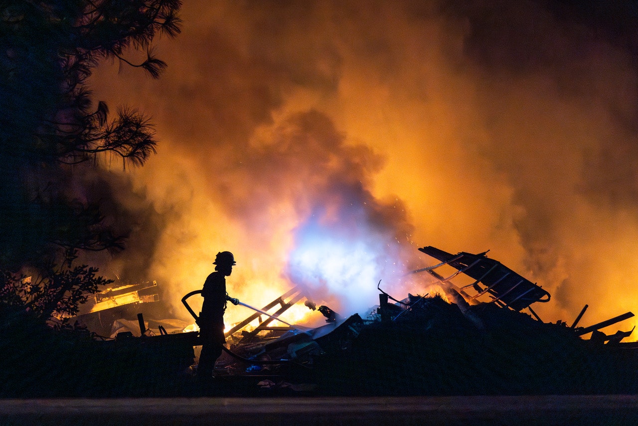 A firefighter silhouetted against a large blaze, with smoke and debris in the background.