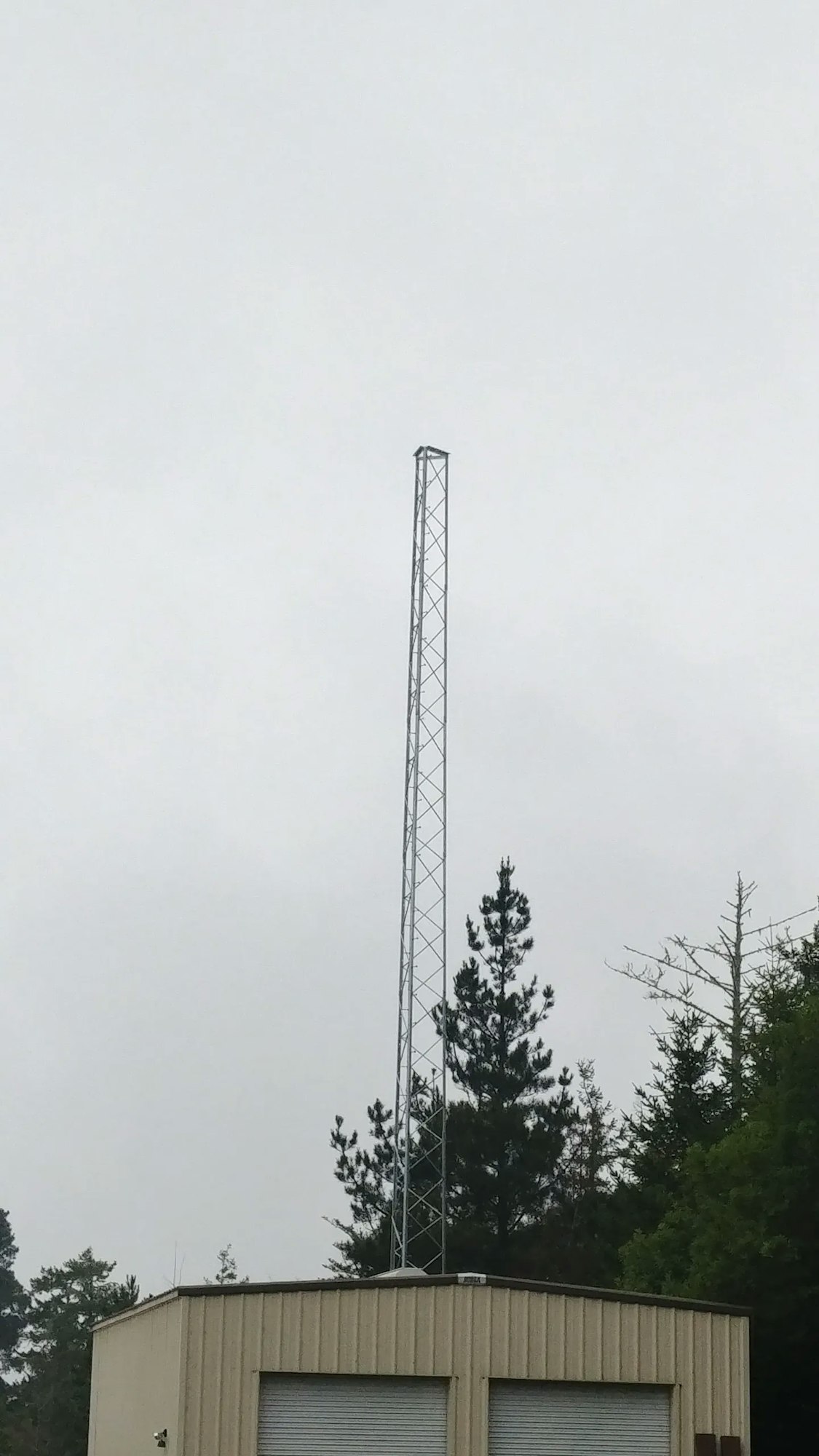 A tall, metal tower structure behind a building with trees under an overcast sky.