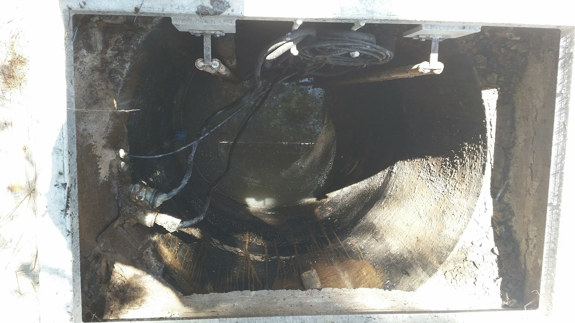 An open manhole revealing underground pipes and cables, with visible water accumulation at the bottom.