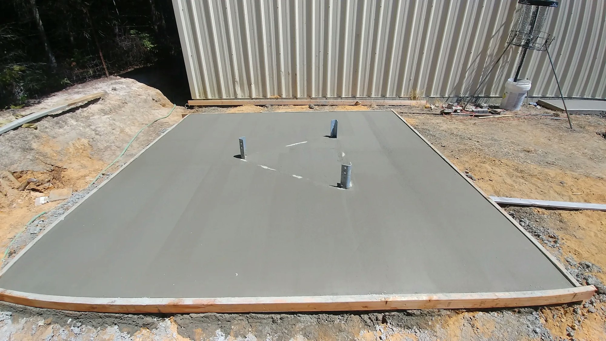 Fresh concrete slab with plumbing protrusions and wooden formwork, near a metal structure and construction materials.
