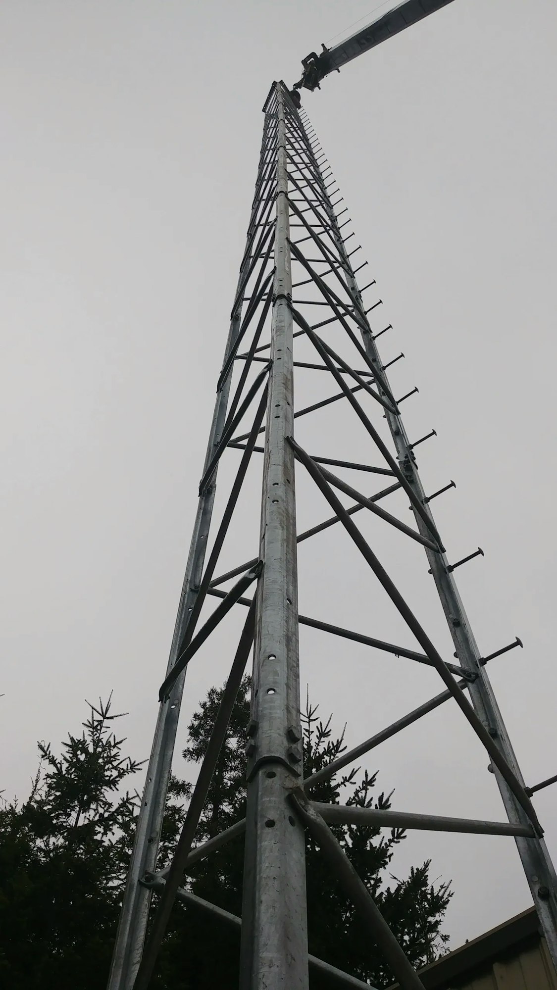 A tall metal tower with a crane extending towards the top, set against a cloudy sky, with trees in the background.