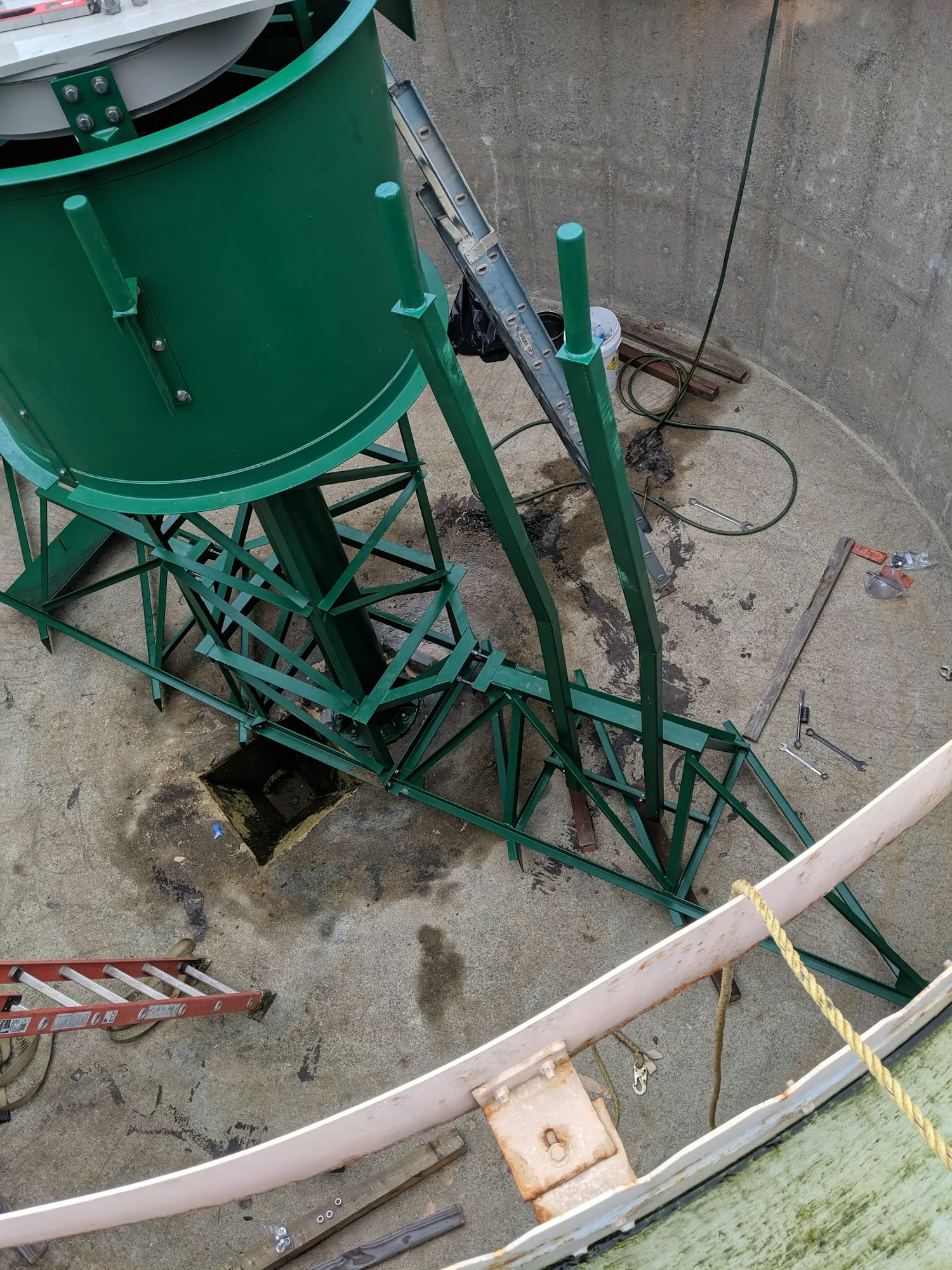 A green silo with a supporting framework, a ladder, construction tools, and safety barriers on a concrete floor.