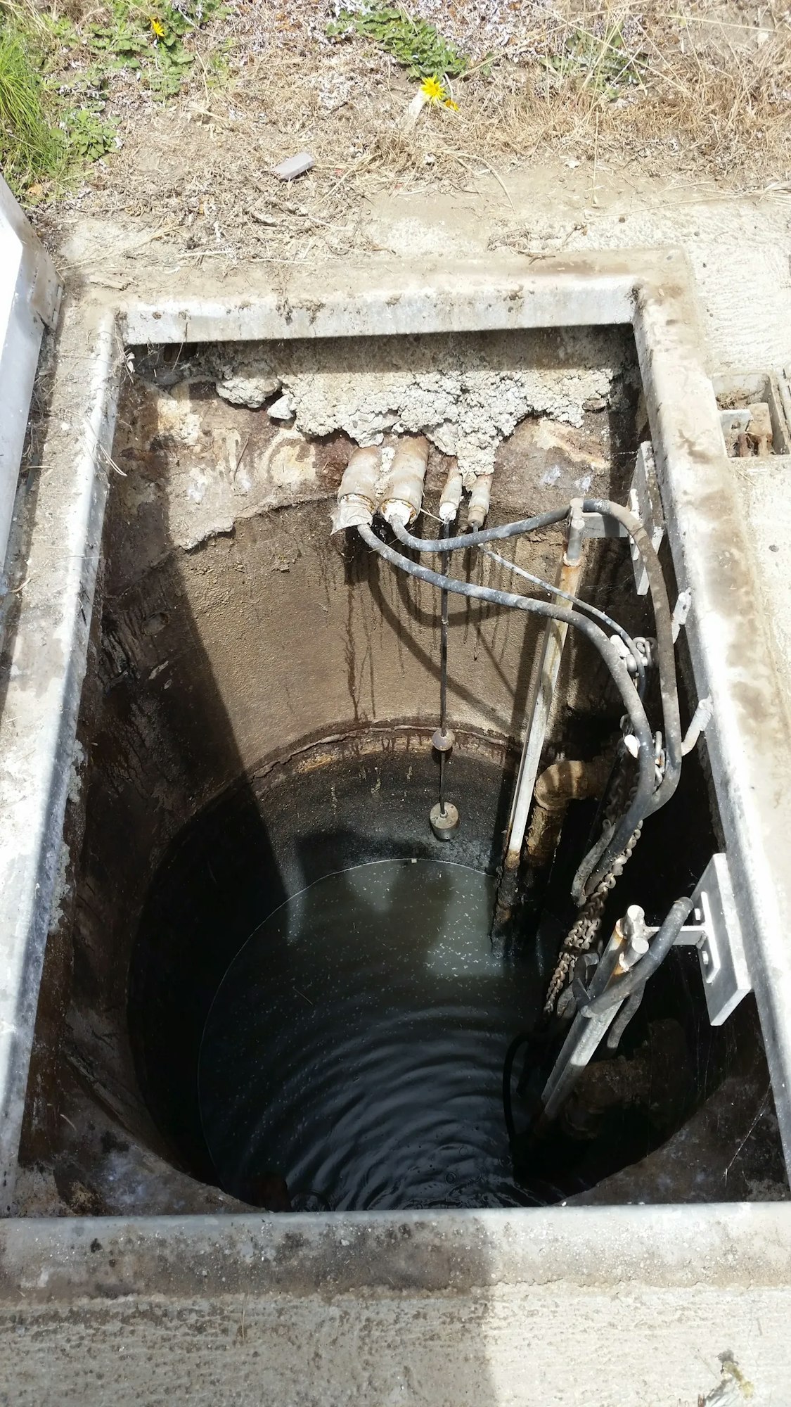 An open manhole with pipes and cables, leading down to water inside.