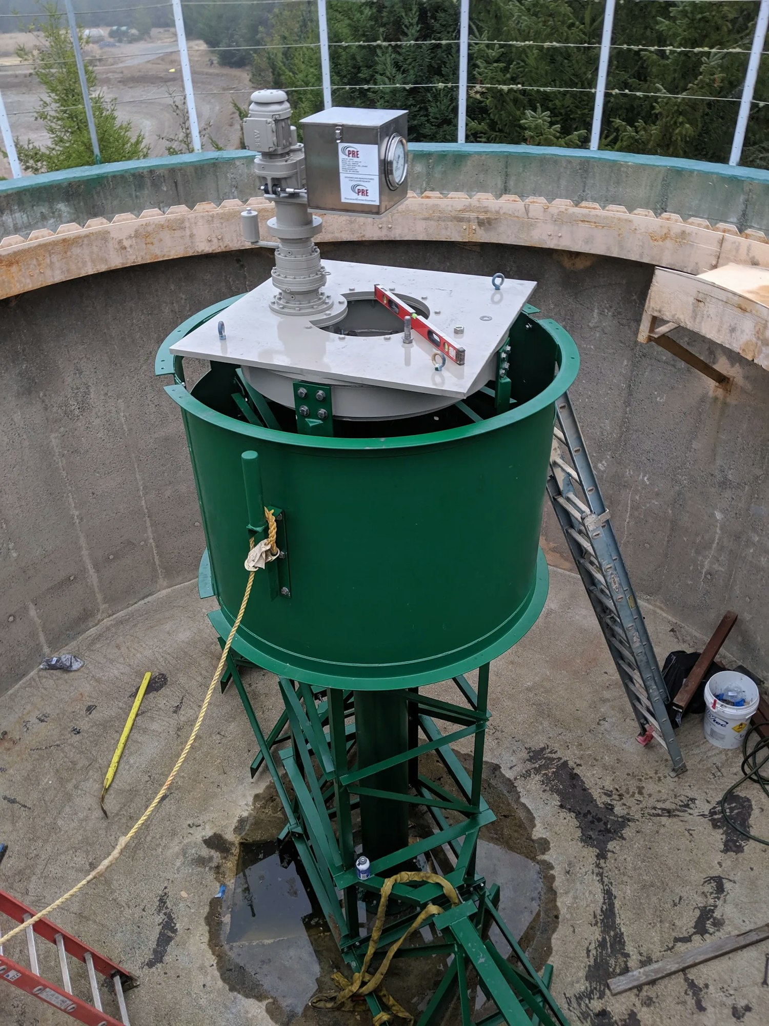 Industrial equipment on a green platform with ladders inside a concrete structure; tools and construction materials scattered around.