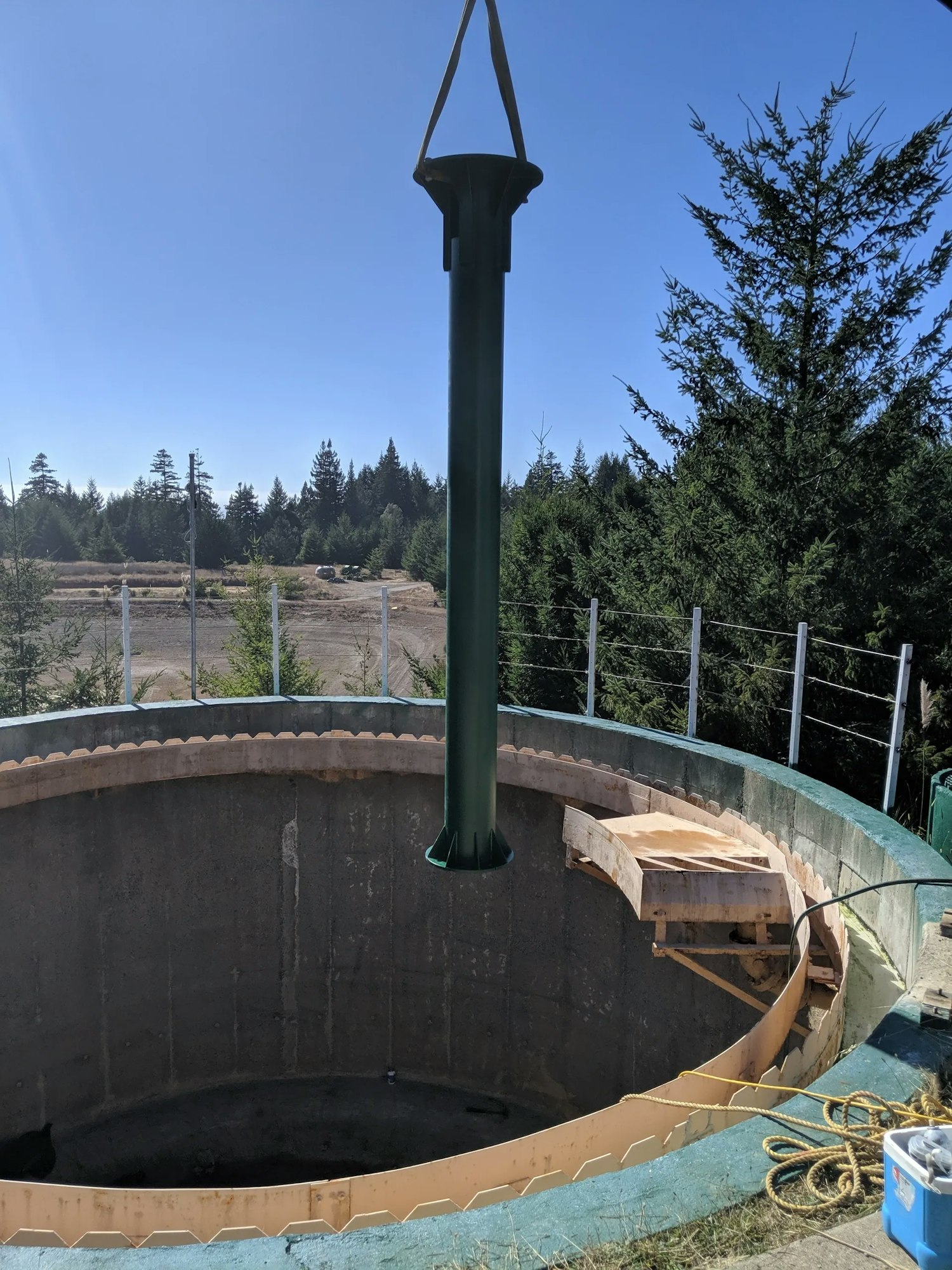 A large, green pipe hanging over a circular concrete structure under construction, with trees and a clear sky in the background.