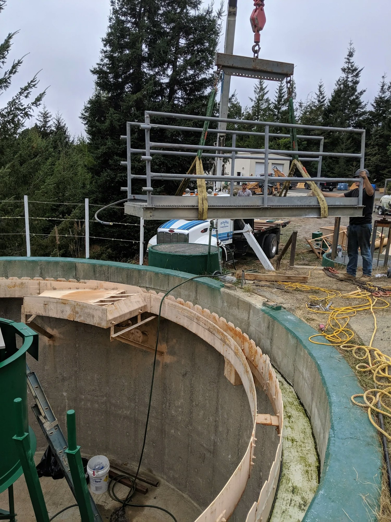 A construction site with a crane lifting a structure, workers, trees in the background, and a circular foundation.