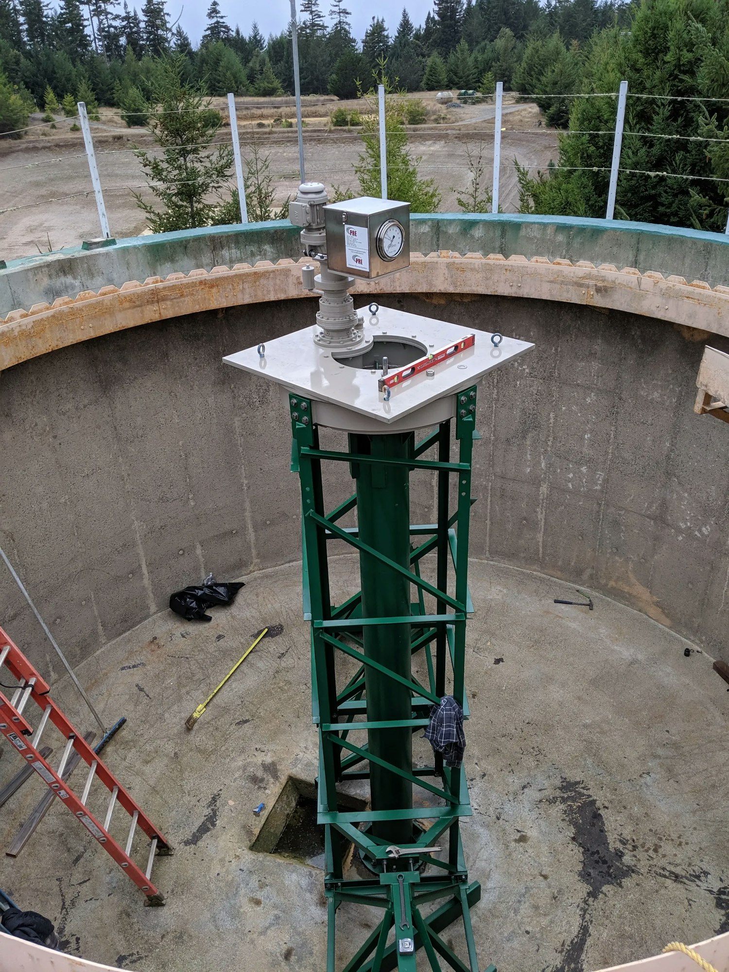 An industrial gauge on a structure inside a large concrete tank with a ladder and tools around, set in a cleared landscape with trees.