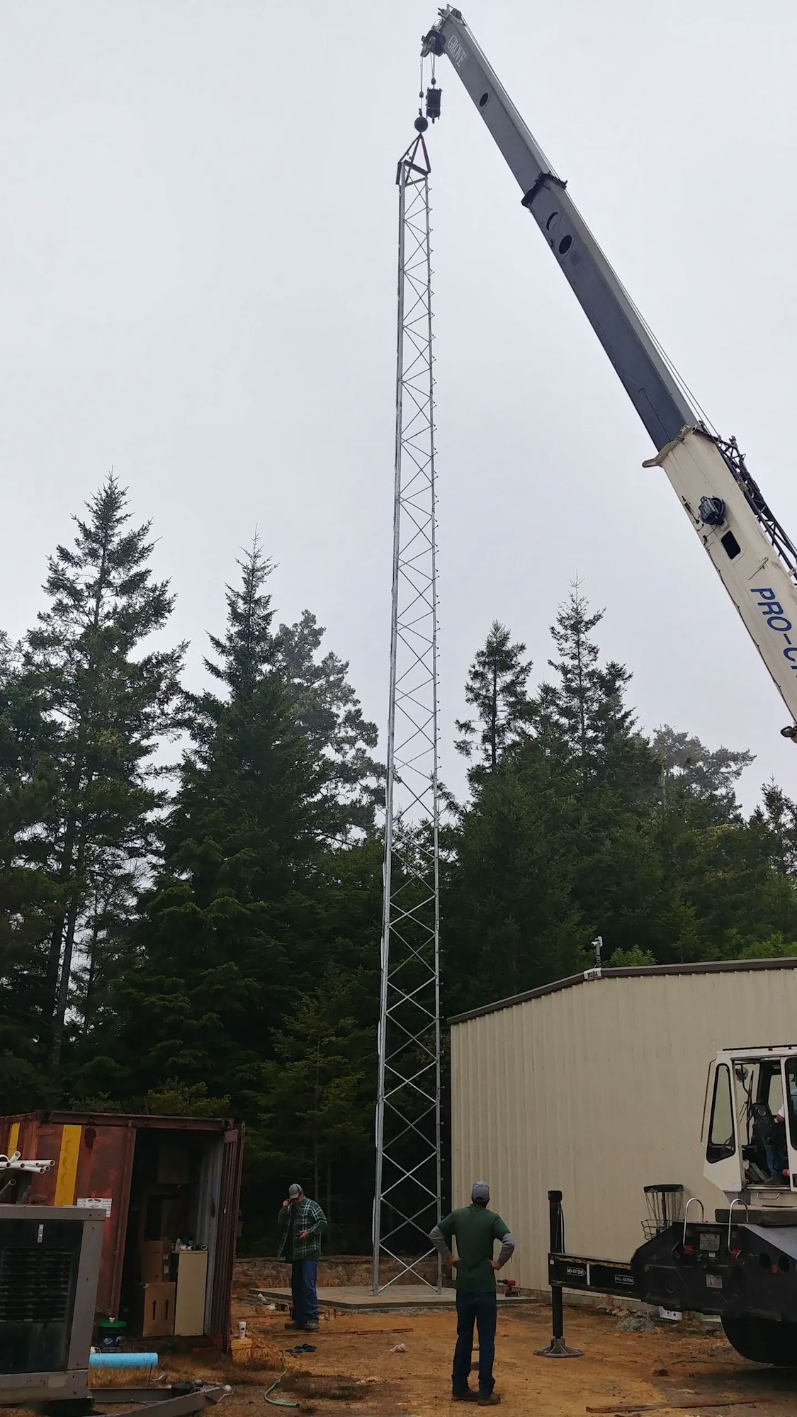 A crane lifting a tall metallic structure, with people overseeing the work, set against a backdrop of trees.