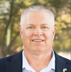 A smiling man in a suit with short hair, standing outdoors with trees in the background.