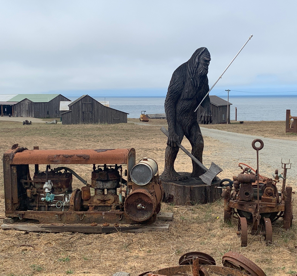 A giant Bigfoot statue stands near rusty machinery and old buildings by the shore, under a cloudy sky.