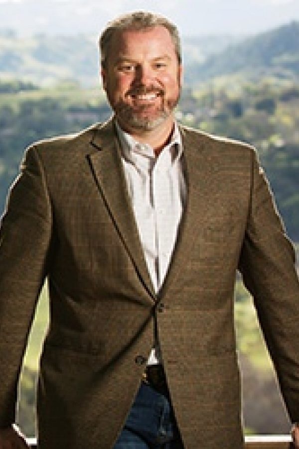 A smiling man in a brown blazer standing outdoors with a scenic background.