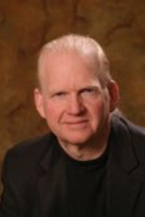 A person with a neutral expression wearing a black top and jacket against a warm, brown background.