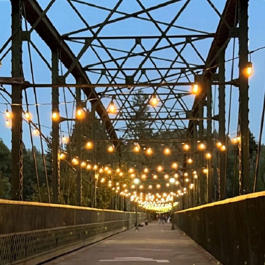 A lit bridge at dusk with string lights and a tree-lined background.