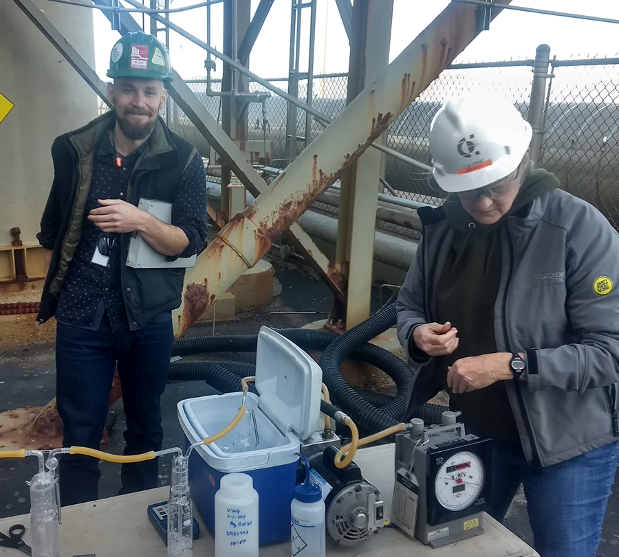 Two people in safety gear are working at an industrial site, focusing on equipment displayed on a table.