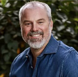 A smiling man with a beard, wearing a denim shirt, is posing in front of green foliage.