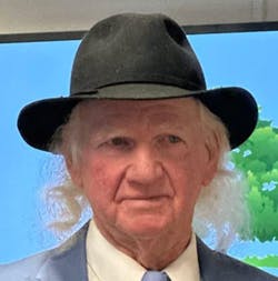 The image shows a man with long hair, wearing a black hat and a suit, against a colorful background.