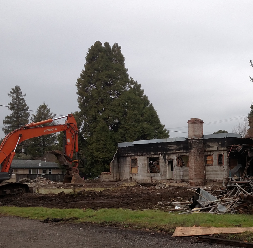 A construction vehicle is demolishing a building, with debris scattered around and tall trees in the background on a cloudy day.