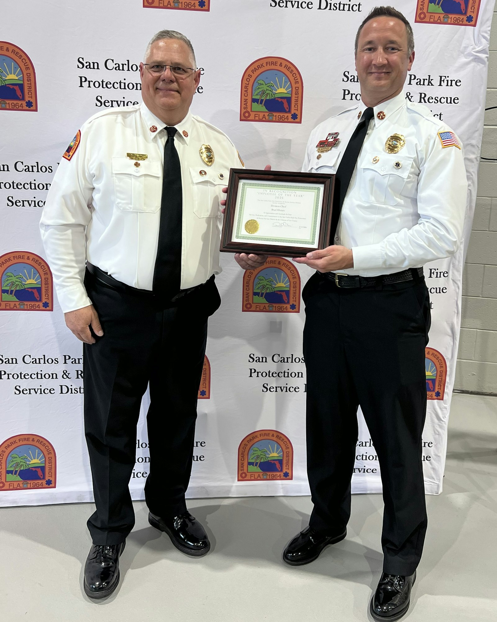 Two uniformed fire service officials are posing with a certificate against a branded backdrop related to the San Carlos Park Fire District.