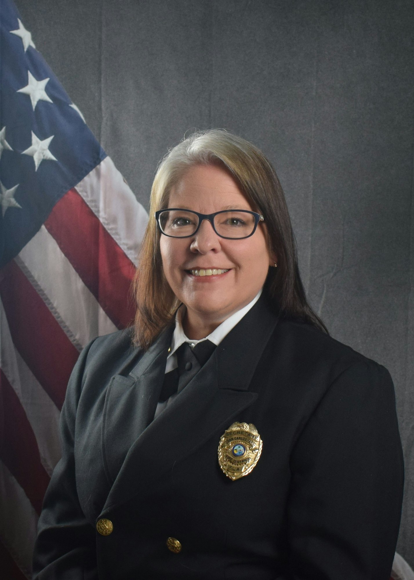 A person in a uniform with glasses, standing in front of a U.S. flag.
