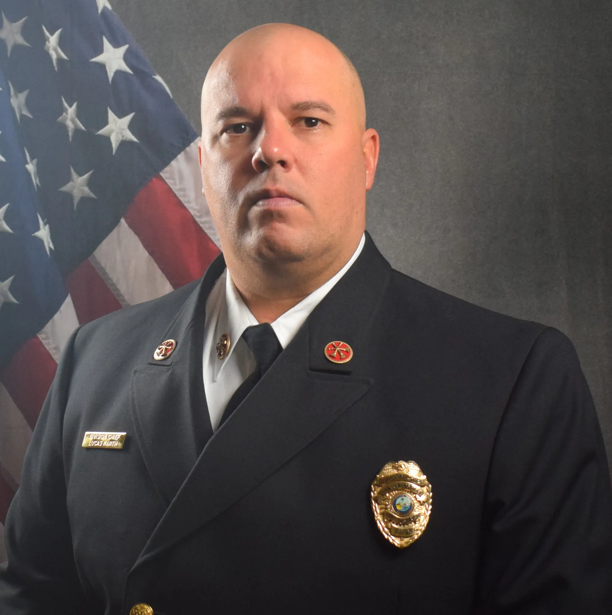 A person in a formal uniform with badges, standing in front of an American flag.
