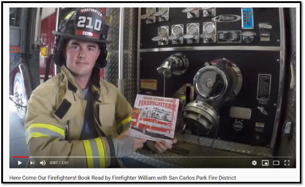 A firefighter holding a book titled "Here Come Our Firefighters!" next to fire truck controls.