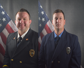 Two men in uniform stand in front of an American flag backdrop.