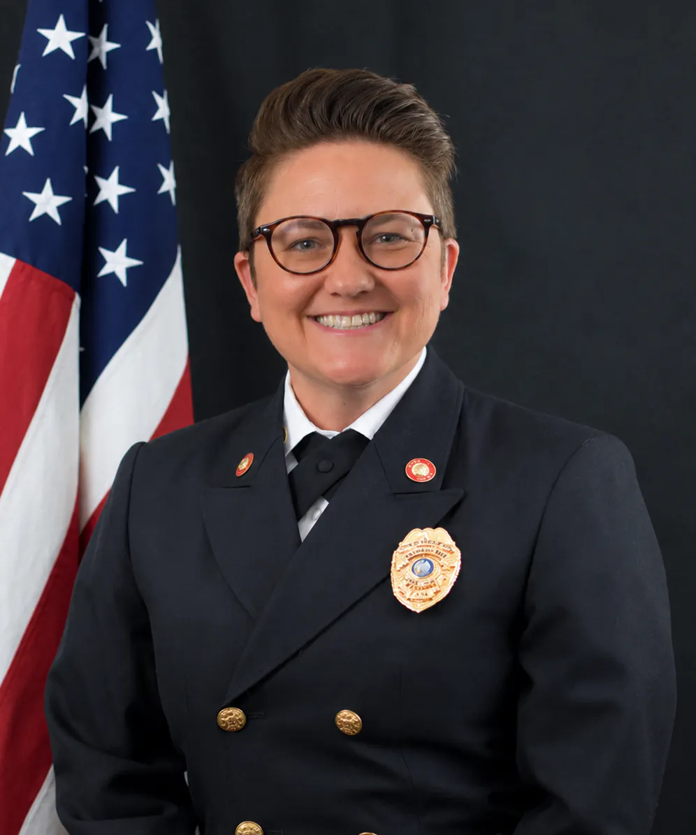 A smiling individual in a formal uniform poses in front of an American flag, showcasing authority and professionalism.