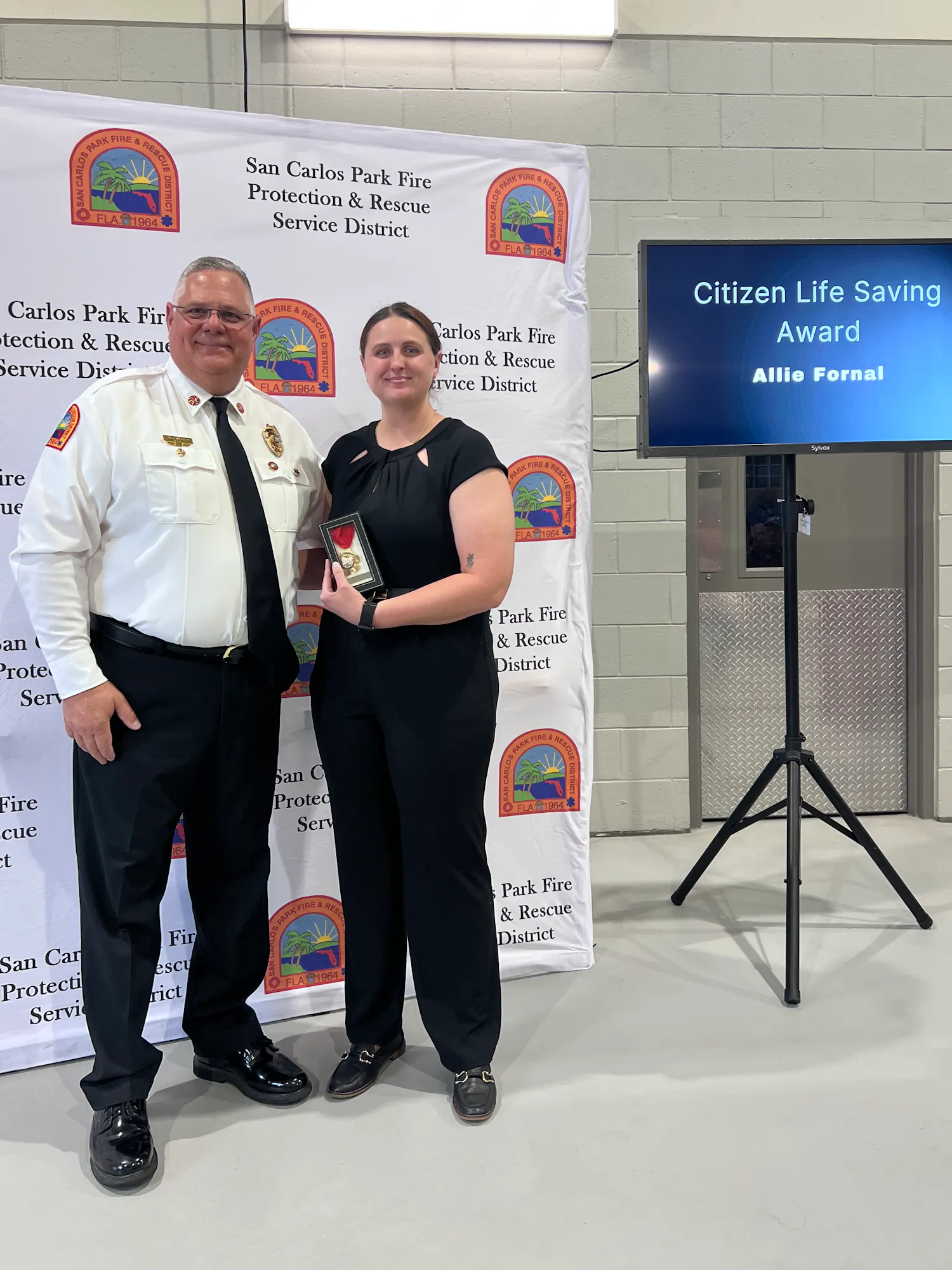 A woman receives a Citizen Life Saving Award from a fire chief at a ceremony, with a backdrop from San Carlos Park Fire District.