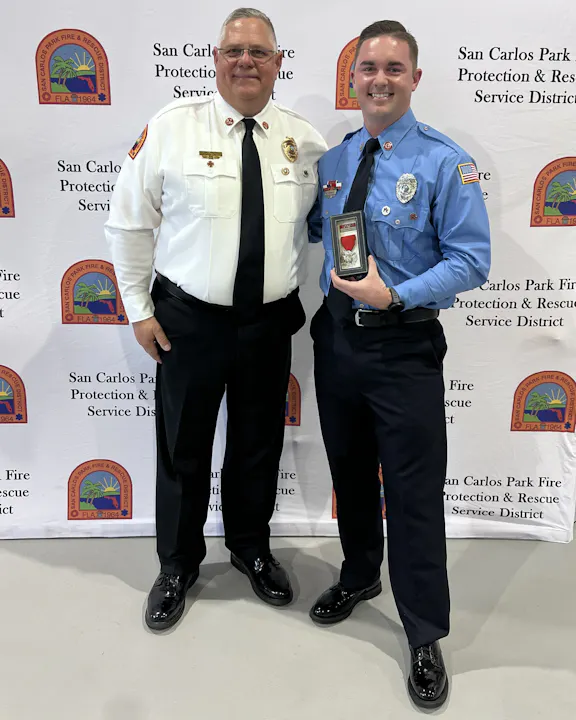 Two men in uniforms stand together, smiling, with a backdrop from the San Carlos Park Fire Protection & Rescue District. One holds an award.