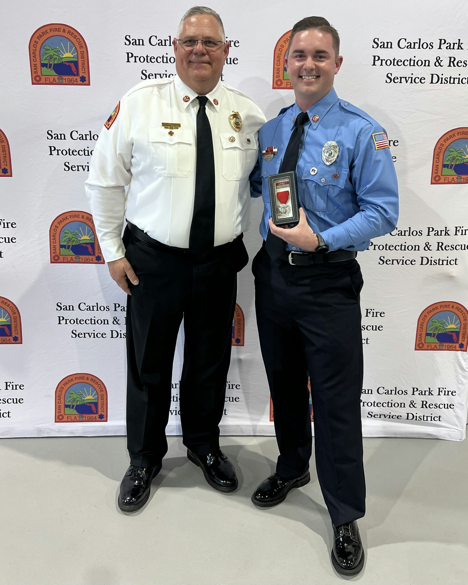 Two men in uniforms stand together, smiling, with a backdrop from the San Carlos Park Fire Protection & Rescue District. One holds an award.
